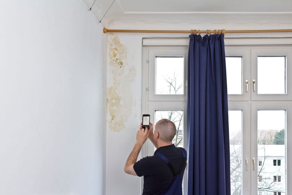 A professional inspecting interior wall water damage caused by a roof leak, capturing photos for documentation and repair planning.